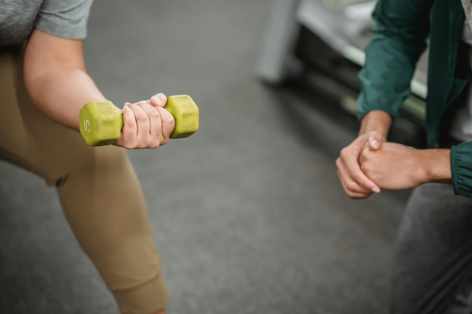 From above of crop anonymous instructor controlling female working out with dumbbell on blurred background of gym