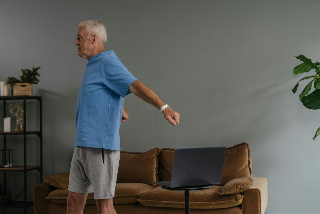 Elderly man stretching indoors while watching laptop, promoting active lifestyle.